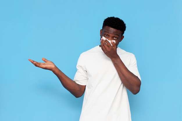 Un joven enfermo afroamericano con camiseta blanca sufre de secreción nasal en el fondo azul