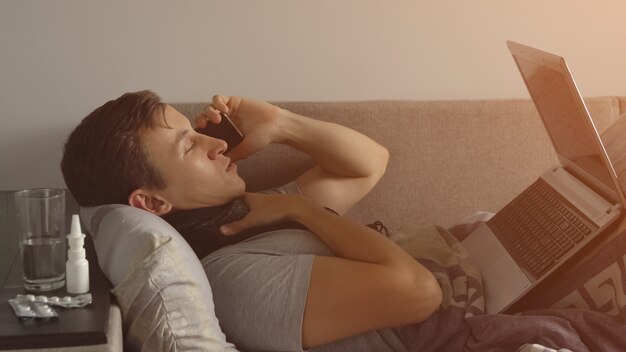 Foto joven enfermo en la cama trabajando en el portátil y hablando por teléfono. hay muchas pastillas y medicamentos en la mesita de noche. luz de sol