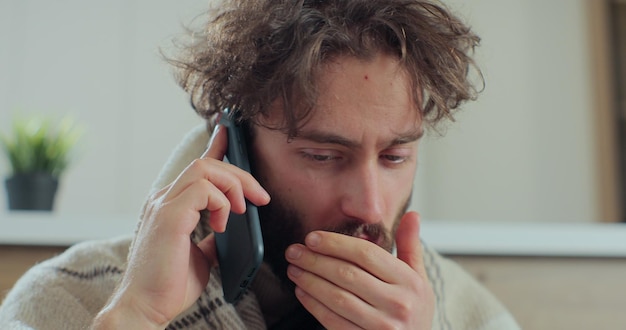 Un joven enfermo hablando por teléfono con un médico mientras se sienta en el sofá, estornudando y tosiendo como si tuviera un resfriado. Concepto de salud y enfermedad.