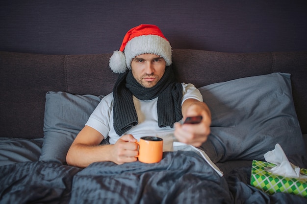 Joven enfermo con sombrero rojo de Navidad se sienta en la cama