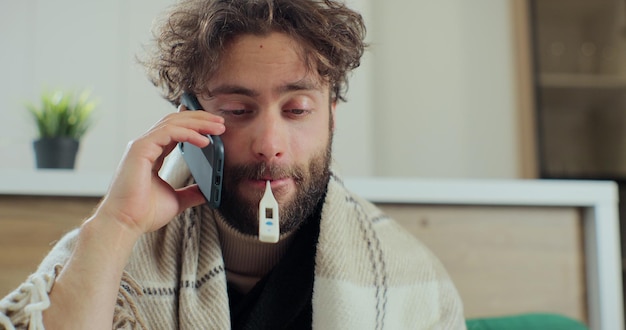 Foto joven enfermo con termómetro en la boca hablando por teléfono mientras se sienta en el sofá, estornudando y tosiendo como si tuviera un resfriado. concepto de salud y enfermedad.