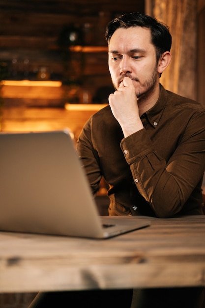 Un joven enfocado con ropa informal escribiendo en una laptop mientras se sienta a la mesa en una acogedora sala de estar. Concepto de trabajo remoto desde casa.