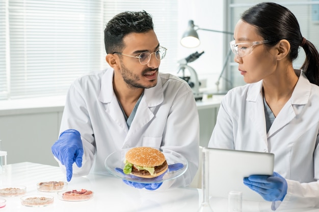 Joven enguantado con hamburguesa en la placa apuntando a una de las muestras de carne vegetal cruda mientras describe sus características a su colega