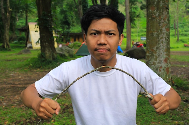 un joven enojado asiático está rompiendo ramas de árboles en el bosque tropical usando una camiseta blanca