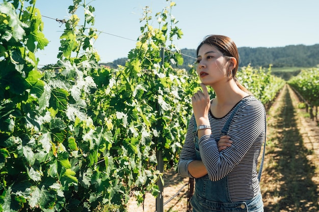 Joven enóloga en viña poniendo las manos debajo de la barbilla mirando los árboles y las plantas pensando en un gran momento. elegante agricultor anfitrión de la bodega de uva concentrado con los brazos cruzados en primavera.