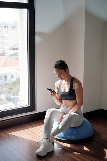 Joven entrenador de fitness asiático sentado en el hemisferio y revisando los mensajes en el teléfono inteligente de sus clientes y colegas