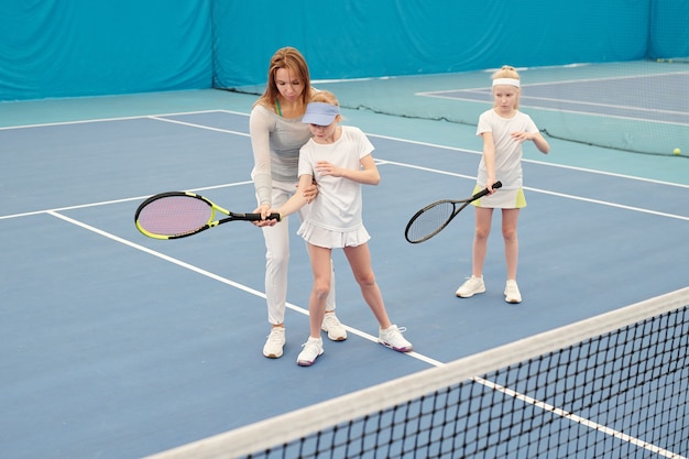 Joven entrenador de tenis en ropa deportiva sosteniendo la raqueta sostenida por una de las dos chicas lindas mientras la consulta durante el entrenamiento individual en el estadio