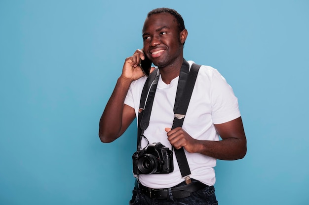 Joven entusiasta de la fotografía confiado con cámara hablando en un moderno dispositivo de teléfono con pantalla táctil. Fotógrafo profesional sonriente hablando por teléfono inteligente mientras está de pie sobre fondo azul.