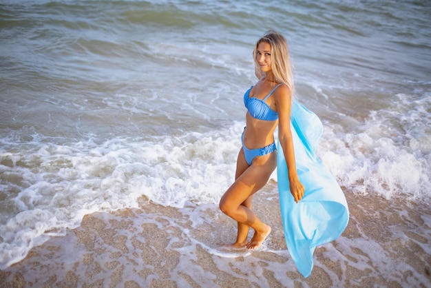 Una joven esbelta y bronceada con cabello rubio en un suave traje de baño azulado y un chal azul claro brillante, camina por la playa cerca del borde del agua de mar disfrutando del cálido verano