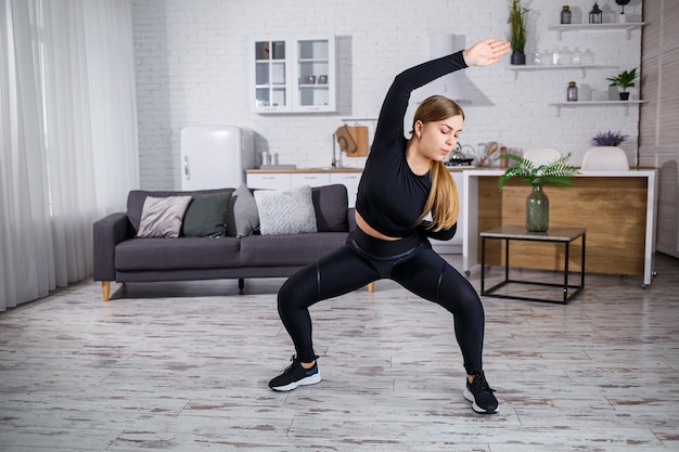 Una joven esbelta practica deportes en casa, está vestida con ropa deportiva, un top negro y leggings. Fitness en casa para un cuerpo hermoso