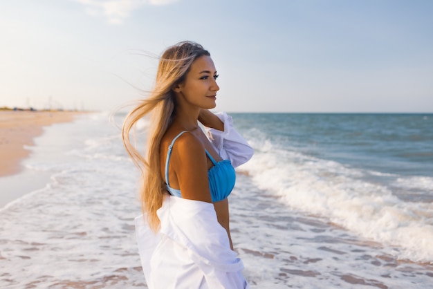 Una joven esbelta en un suave traje de baño azul y una camisa blanca, camina por una amplia playa de arena cerca del mar azul con olas blancas.