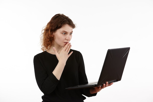 Una joven esbelta vestida de negro trabaja en una computadora portátil. Figura aislada sobre fondo blanco.