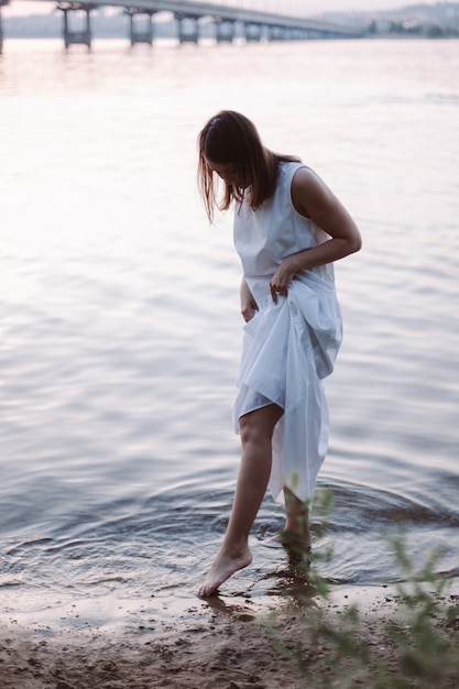 Una joven esbelta con un vestido blanco de verano que se levanta el dobladillo de su vestido moja sus pies en el agua ...