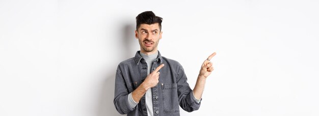 Joven escéptico y dudoso señalando con el dedo a la izquierda el aspecto del logotipo con aversión y confusión