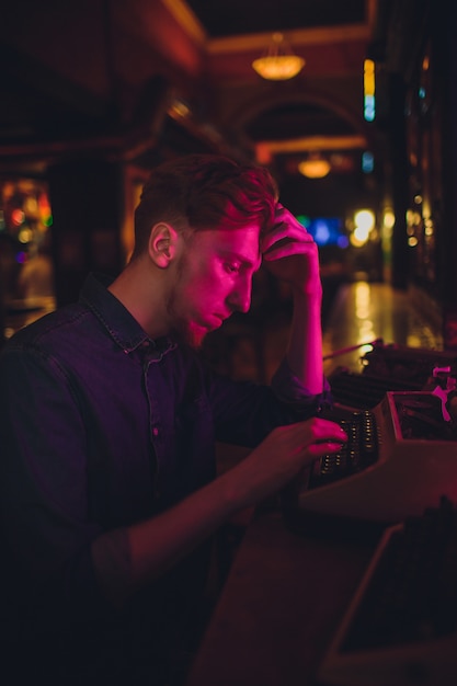 Joven escribiendo en una vieja máquina de escribir. en iluminación oscura, restaurante, ropa moderna, viejos hábitos de escritor