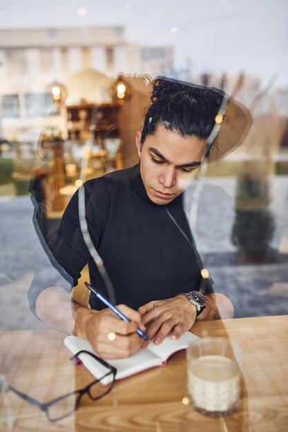 Joven escritor masculino con cabello rizado negro sentado en el interior de un café con bolígrafo y bloc de notas. Ver a través del cristal.