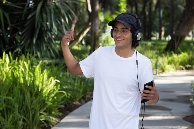 Joven escuchando música con auriculares mientras pasea por el parque sonriendo y relajado en medio de la naturaleza, con gorra y camiseta blanca