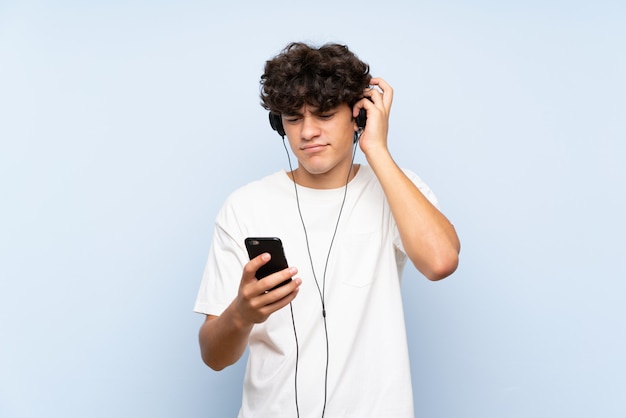 Joven escuchando música con un móvil sobre pared azul aislado