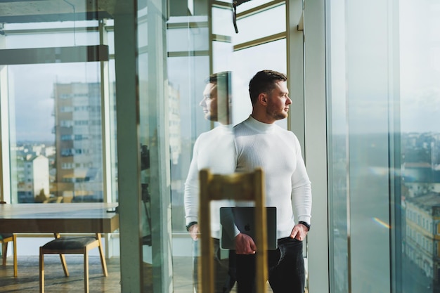 Un joven en un espacio de trabajo moderno se encuentra cerca de una ventana grande Un hombre vestido con ropa informal con una computadora portátil en las manos Trabajo remoto Buen joven gerente