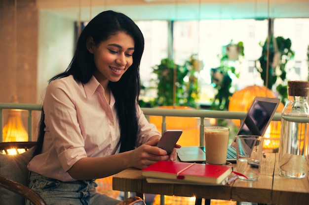 Joven especialista trabajando en la mesa de café con una computadora portátil y sonriendo mientras mira la pantalla de su teléfono inteligente