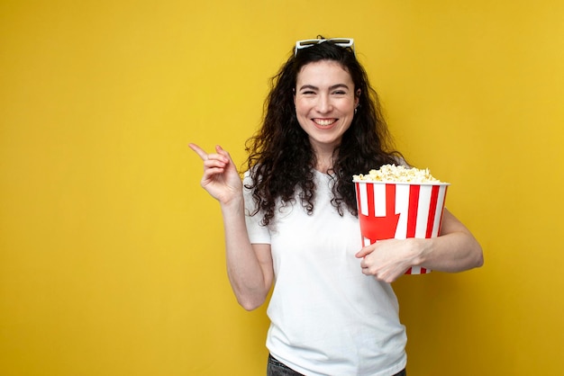 Una joven espectadora morena con gafas 3D y camiseta blanca sostiene un gran cubo de palomitas de maíz