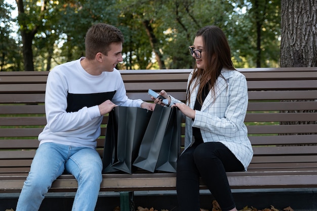 Joven esposo y esposa están sosteniendo bolsas de la compra y están considerando ir de compras en un banco en el parque.