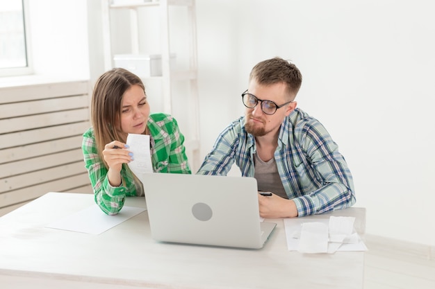 Un joven esposo y una esposa insatisfechos cuentan las facturas de servicios públicos para el pago de un apartamento y escriben los resultados en un cuaderno y una computadora portátil en la contabilidad de su hogar.