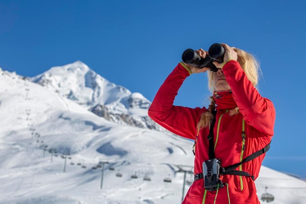Una joven esquiadora con traje rojo mira a través de binoculares a las altas montañas
