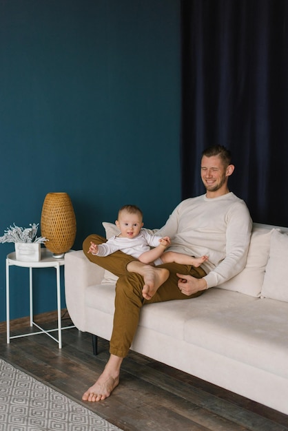 El joven está feliz y sostiene a su hijo de seis meses en sus brazos sentado en el sofá de la habitación.