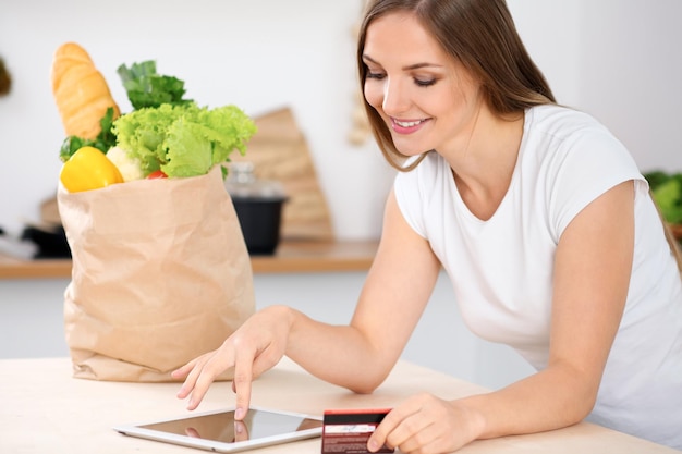 Una joven está haciendo compras en línea con una tableta y una tarjeta de crédito. El ama de casa encontró una nueva receta para cocinar en la cocina. Una gran bolsa de papel llena de verduras y frutas frescas está parada en la mesa.