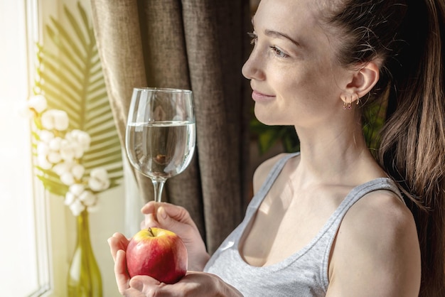 Una joven está parada frente a la ventana en una mañana soleada y sosteniendo un vaso de agua clara y una manzana en sus manos Concepto de un comienzo agradable del día y hábitos saludables