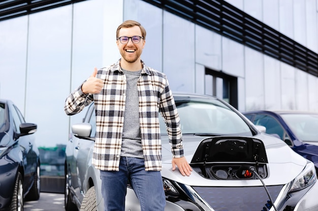 Un joven está de pie junto a su nuevo coche eléctrico y descansa mientras el coche se carga