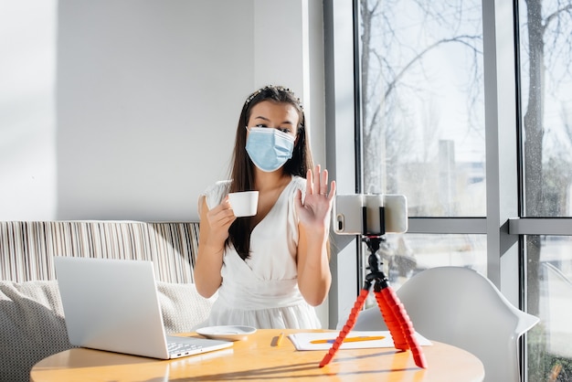 Una joven está sentada en un café con una máscara y dirige un blog de video. Comunicación a la cámara.
