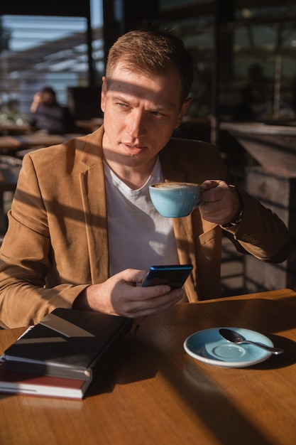 un joven está sentado en un café esperando una reunión de negocios tomando café y sosteniendo un teléfono