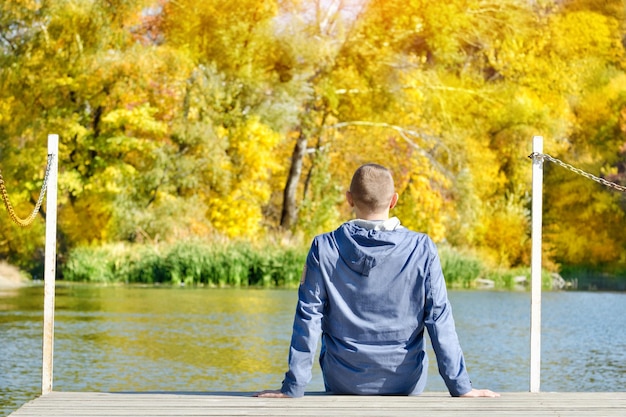 Joven está sentado en el muelle Otoño soleado Vista posterior