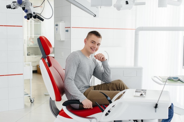 Un joven está sentado en una silla dental roja y sonriendo en odontología blanca moderna Tratamiento y prevención de caries desde la juventud Odontología moderna y prótesis