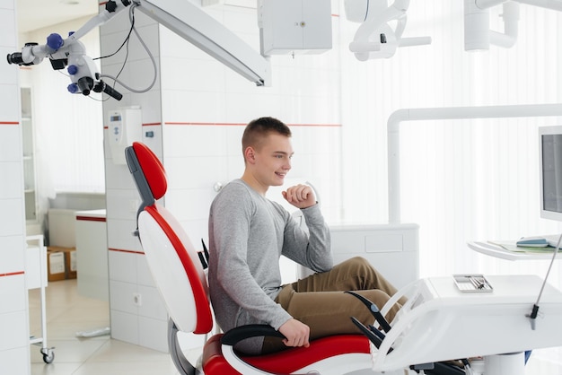 Un joven está sentado en una silla dental roja y sonriendo en odontología blanca moderna Tratamiento y prevención de caries desde la juventud Odontología moderna y prótesis