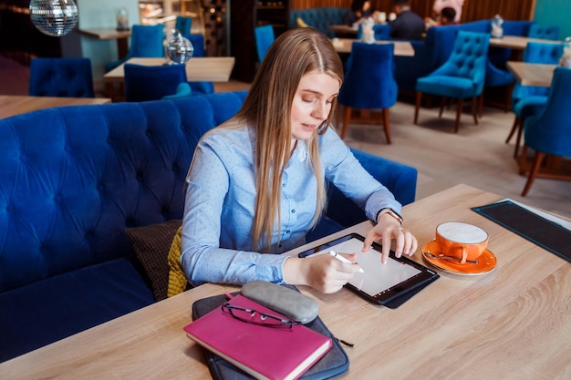 La joven está trabajando en su tableta en la taza de café junto a ella en la mesa