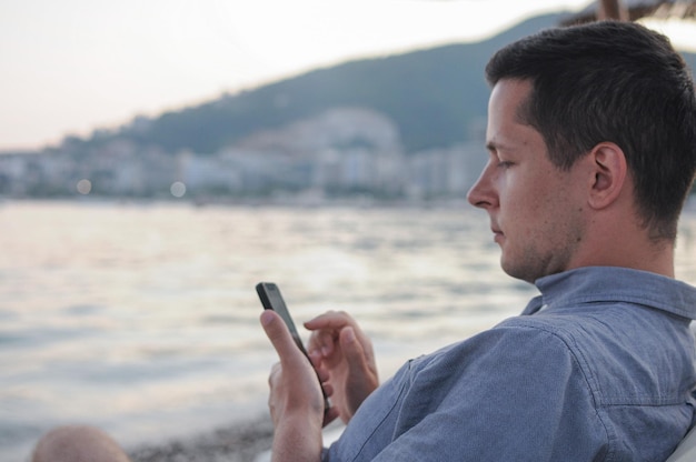 Un joven está usando su teléfono móvil mientras está sentado en la playa cerca del mar