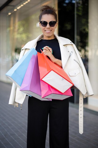 Una joven estadounidense feliz con una camiseta negra, una chaqueta blanca y gafas de sol con bolsas coloridas