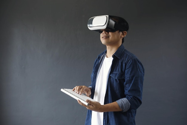 Joven de estilo casual usando gafas de realidad virtual y teclado inalámbrico en un fondo gris oscuro