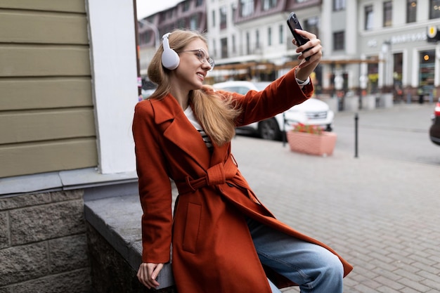 Una joven estudiante con un abrigo rojo y auriculares se toma una selfie con el telón de fondo de la ciudad