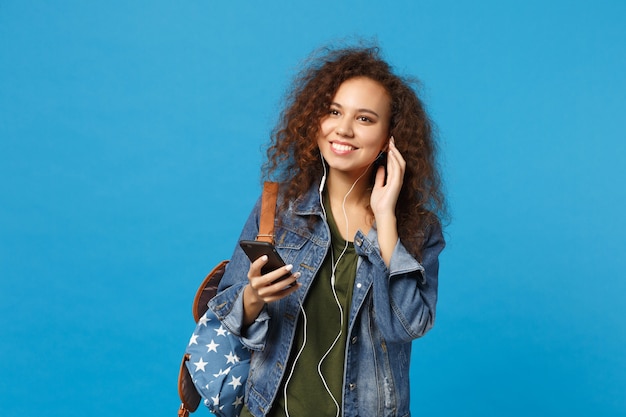 Joven estudiante adolescente afroamericana en ropa de mezclilla, auriculares de mochila aislado en la pared azul