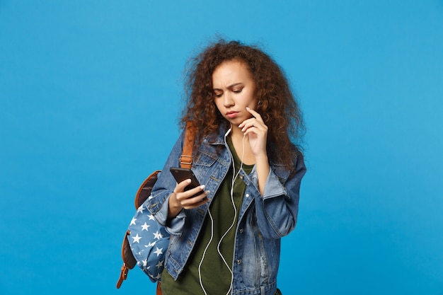 Joven estudiante adolescente afroamericana en ropa de mezclilla, auriculares de mochila aislado en la pared azul