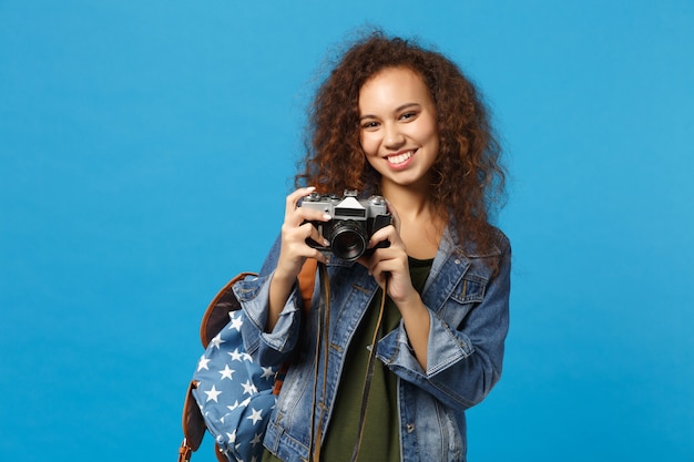 Joven estudiante adolescente afroamericana en ropa de mezclilla mochila mantenga cámara aislada en la pared azul