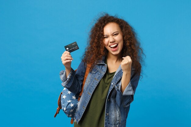 Foto joven estudiante adolescente afroamericana en ropa de mezclilla, mochila mantenga tarjeta de crédito aislada en la pared azul