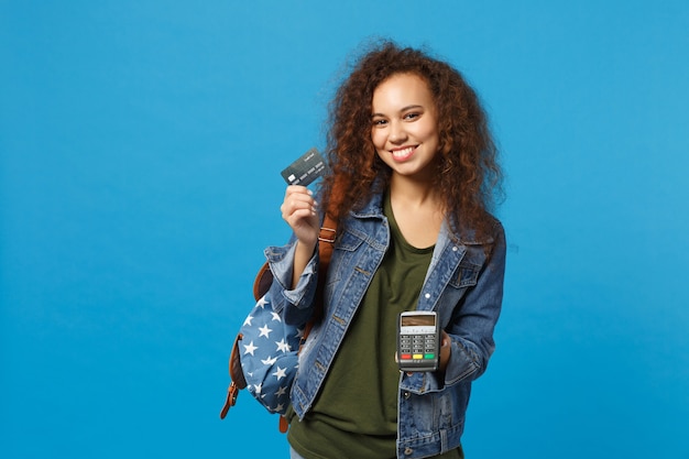 Joven estudiante adolescente afroamericana en ropa de mezclilla, mochila mantenga tarjeta de crédito aislada en la pared azul