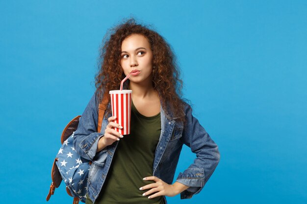 Joven estudiante adolescente afroamericana en ropa de mezclilla, mochila mantenga taza de papel aislada en la pared azul