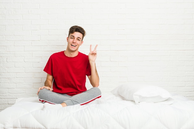 Joven estudiante adolescente hombre en la cama alegre y despreocupado mostrando un símbolo de paz con los dedos.