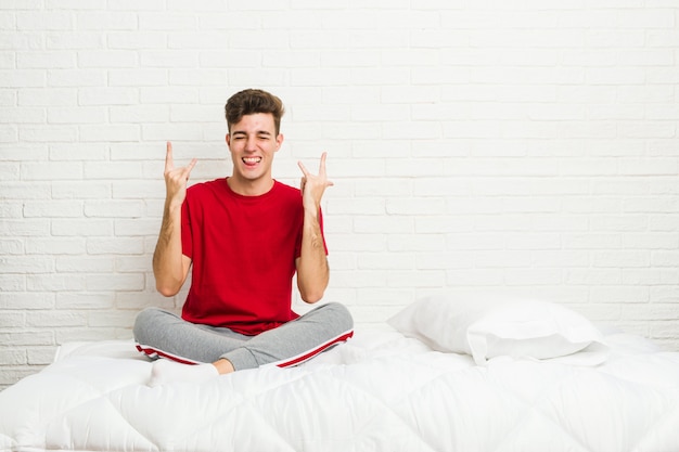 Joven estudiante adolescente hombre en la cama mostrando el gesto de rock con los dedos